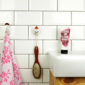 May include: White bathroom with white subway tile walls. A pink floral hand towel hangs on a ceramic hook. A wooden hairbrush hangs on a ceramic hook shaped like a mushroom. A white porcelain sink with a chrome faucet. A bottle of shower gel with the text "Loved Up" on the front.