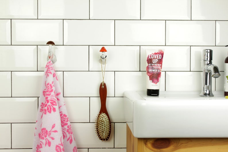 May include: White bathroom with white subway tile walls. A pink floral hand towel hangs on a ceramic hook. A wooden hairbrush hangs on a ceramic hook shaped like a mushroom. A white porcelain sink with a chrome faucet. A bottle of shower gel with the text "Loved Up" on the front.