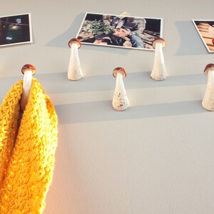 May include: White ceramic mushroom-shaped hooks with brown tops mounted on a wall. A yellow knitted scarf is hanging from one of the hooks. There are three framed photographs of couples on the wall.