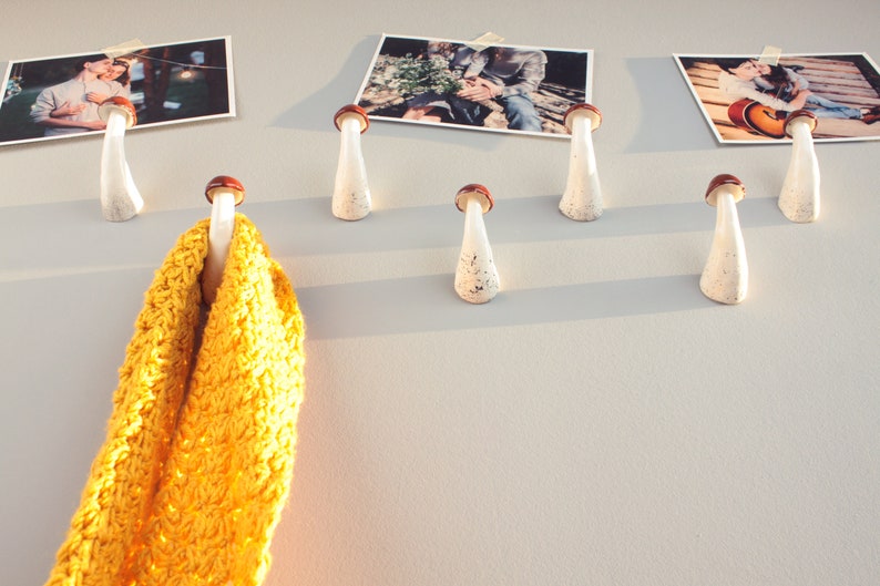 May include: White ceramic mushroom-shaped hooks with brown tops mounted on a wall. A yellow knitted scarf is hanging from one of the hooks. There are three framed photographs of couples on the wall.