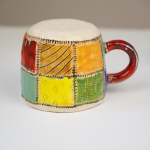 K&ouml;nnte beinhalten: Eine Keramik-Kaffeetasse mit Patchwork-Design in Rot-, Orange-, Gelb-, Gr&uuml;n- und Blaut&ouml;nen. Die Tasse hat ein wei&szlig;es Innenleben und einen roten Henkel.