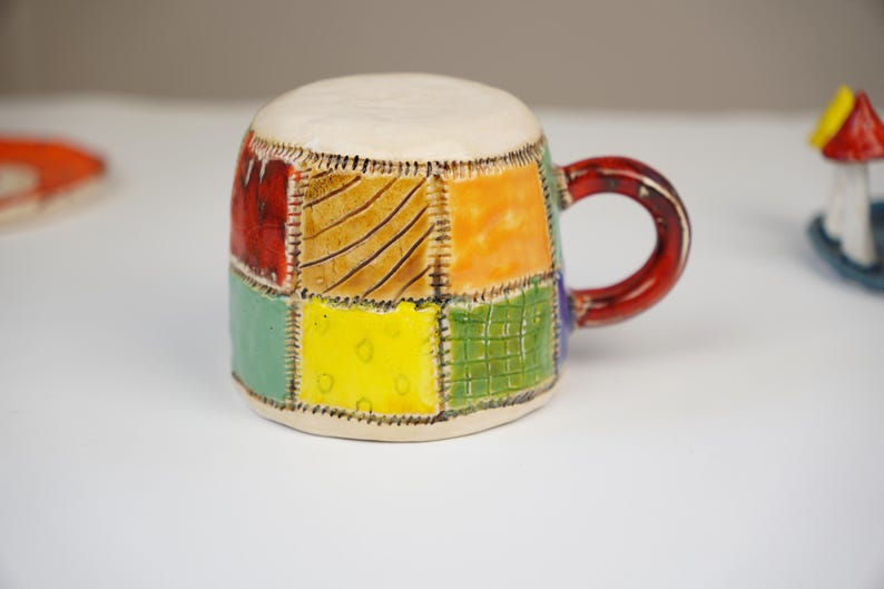 K&ouml;nnte beinhalten: Eine Keramik-Kaffeetasse mit Patchwork-Design in Rot-, Orange-, Gelb-, Gr&uuml;n- und Blaut&ouml;nen. Die Tasse hat ein wei&szlig;es Innenleben und einen roten Henkel.
