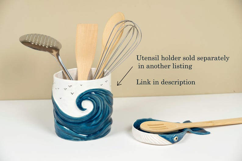 May include: A white ceramic utensil holder shaped like a wave with blue and white accents. The holder is filled with kitchen utensils, including a wooden spoon, a whisk, and a slotted spoon.  The text "Utensil holder sold separately in another listing. Link in description" is visible in the image.