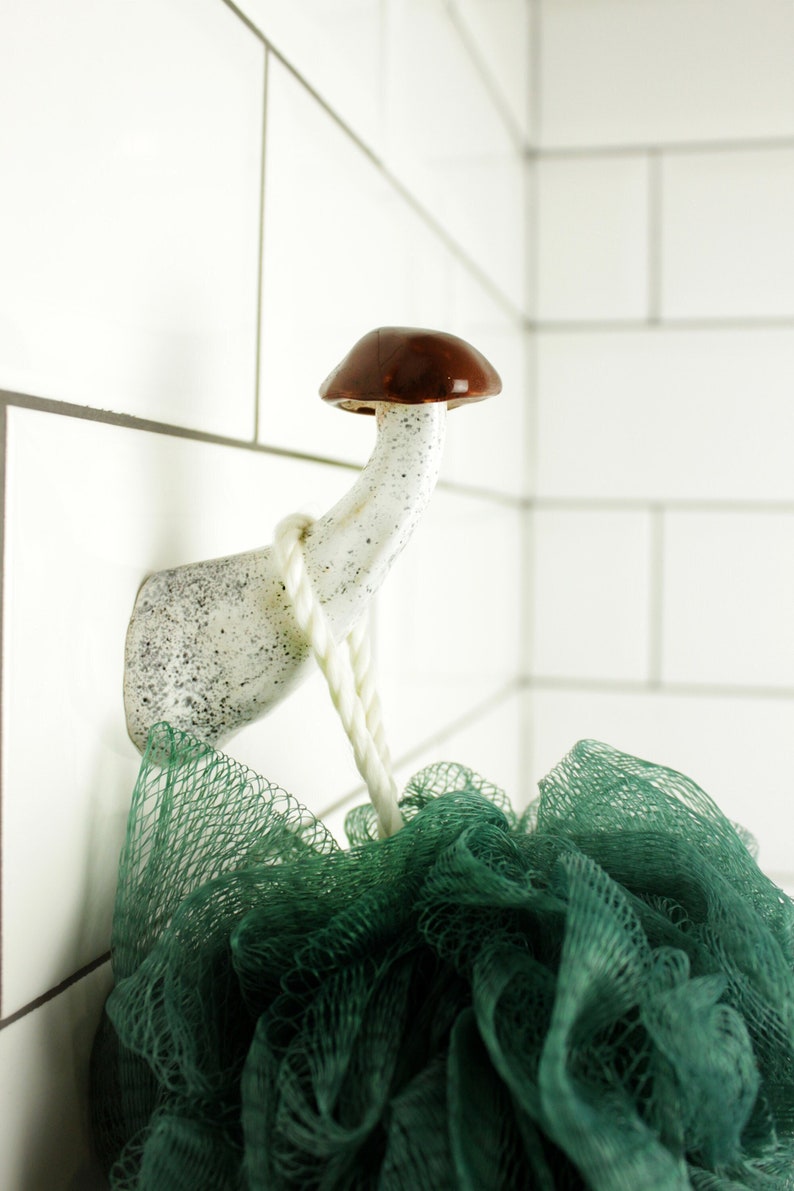 May include: A white ceramic mushroom-shaped hook with a brown top, hanging on a white tiled wall. A green bath pouf is hanging from the hook.