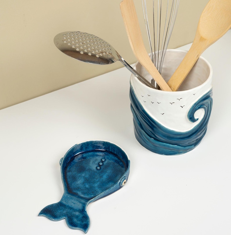 May include: A blue and white ceramic utensil holder shaped like a wave with seagulls flying above. The holder is filled with kitchen utensils, including a wooden spoon, a whisk, and a slotted spoon. A matching blue ceramic spoon rest shaped like a whale is on the counter.