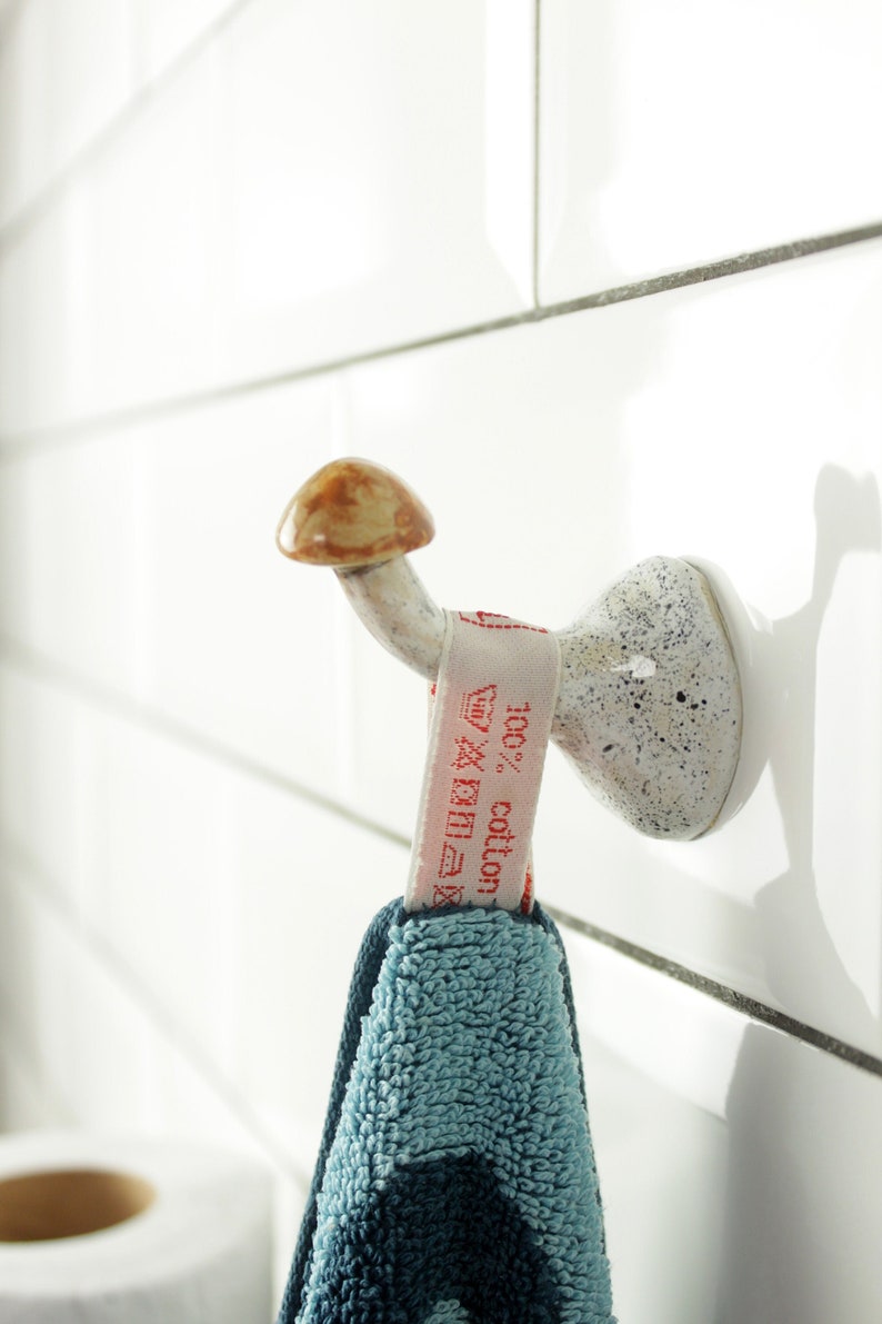 May include: A white ceramic mushroom-shaped hook with a brown top, mounted on a white tiled wall. A blue and green towel with a white tag that reads "100% cotton" is hanging from the hook.