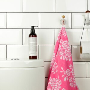 May include: A white bathroom with white subway tiles, a white toilet, a pink hand towel hanging on a mushroom-shaped hook, and a roll of toilet paper hanging on a mushroom-shaped hook.