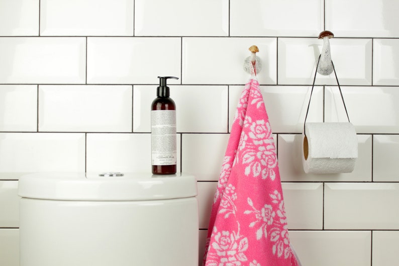 May include: A white bathroom with white subway tiles, a white toilet, a pink hand towel hanging on a mushroom-shaped hook, and a roll of toilet paper hanging on a mushroom-shaped hook.
