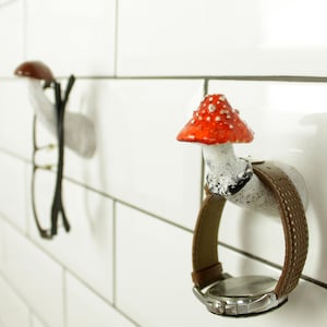 May include: Two ceramic mushroom-shaped hooks mounted on a white tiled wall. One hook has a red and white mushroom cap and holds a brown leather wristwatch. The other hook has a brown mushroom cap and holds a pair of black eyeglasses.