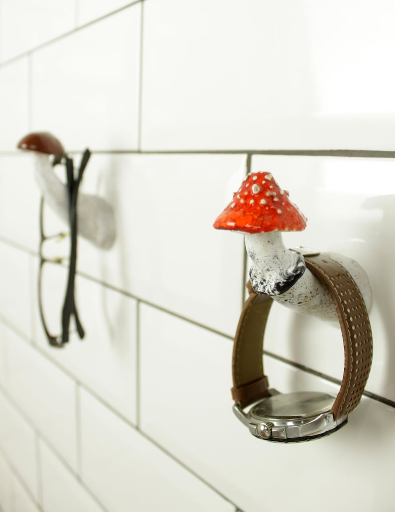 May include: Two ceramic mushroom-shaped hooks mounted on a white tiled wall. One hook has a red and white mushroom cap and holds a brown leather wristwatch. The other hook has a brown mushroom cap and holds a pair of black eyeglasses.