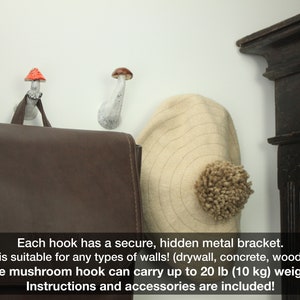 May include: Two white ceramic mushroom hooks with red and brown tops mounted on a white wall. A brown leather bag hangs from one hook and a beige hat hangs from the other. The text below reads: "Each hook has a secure, hidden metal bracket. The mount is suitable for any types of walls! (drywall, concrete, wood and other!) The mushroom hook can carry up to 20 lb (10 kg) weight! Instructions and accessories are included!"