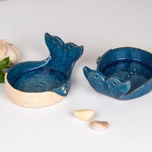 May include: Two ceramic whale-shaped dishes with a glossy blue glaze. The dishes have a cream-colored base and are decorated with carved details. A bulb of garlic, basil leaves, and garlic cloves are next to the dishes.