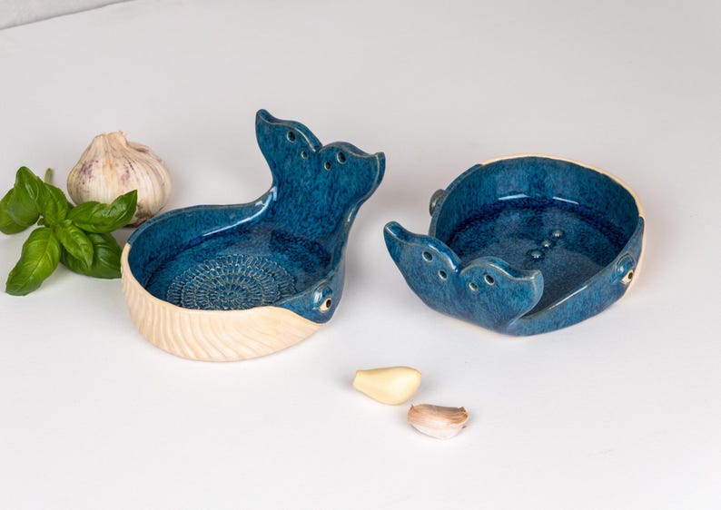 May include: Two ceramic whale-shaped dishes with a glossy blue glaze. The dishes have a cream-colored base and are decorated with carved details. A bulb of garlic, basil leaves, and garlic cloves are next to the dishes.