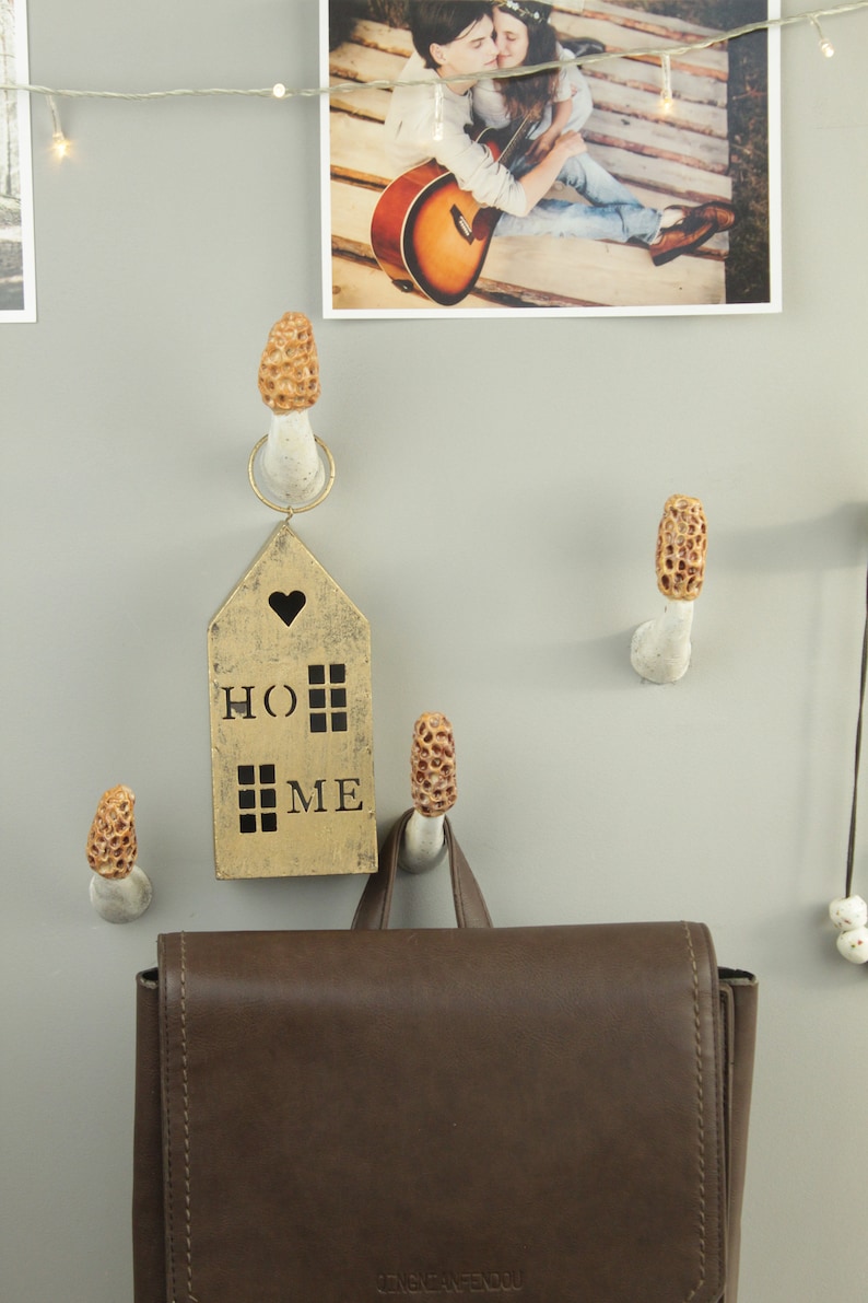 May include: A brown leather backpack hangs on a wall with a gold house-shaped sign that says "HOME" with a heart above the "O".  The house sign is hanging from a white ceramic mushroom hook.  There are three other mushroom hooks on the wall.
