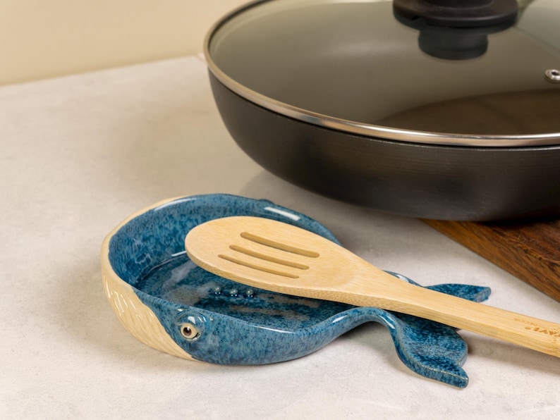 May include: A blue ceramic whale-shaped spoon rest with a wooden spoon resting inside. The spoon rest has a white rim and a speckled blue glaze.