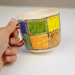 K&ouml;nnte beinhalten: Eine bunte Keramiktasse mit Patchwork-Design. Die Tasse hat einen roten Henkel und ist mit Quadraten in Gr&uuml;n, Gelb, Orange, Blau und Braun verziert.