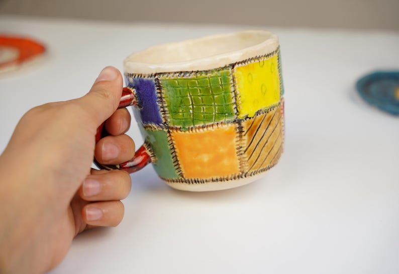 K&ouml;nnte beinhalten: Eine bunte Keramiktasse mit Patchwork-Design. Die Tasse hat einen roten Henkel und ist mit Quadraten in Gr&uuml;n, Gelb, Orange, Blau und Braun verziert.