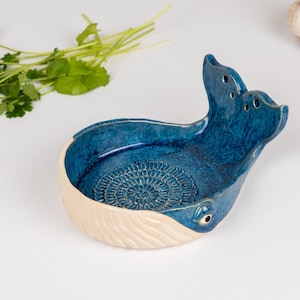 May include: A ceramic whale-shaped garlic grater in shades of blue and cream. The grater has a textured interior and a tail with holes. Fresh cilantro and garlic cloves are also visible.