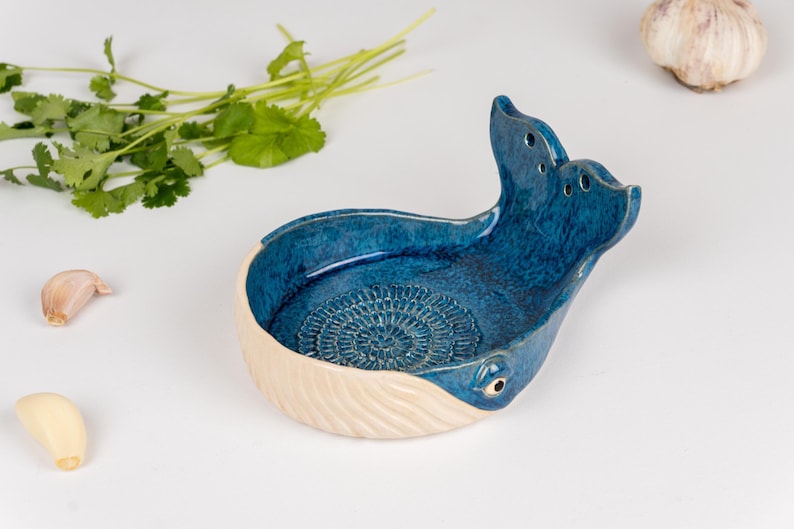 May include: A ceramic whale-shaped garlic grater in shades of blue and cream. The grater has a textured interior and a tail with holes. Fresh cilantro and garlic cloves are also visible.
