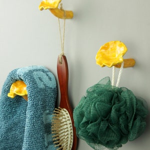 May include: Two yellow ceramic mushroom-shaped hooks mounted on a wall. A blue towel is hanging on one hook, a wooden hairbrush is hanging on the other, and a green bath sponge is hanging on the bottom of the second hook.
