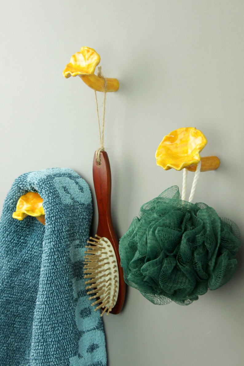 May include: Two yellow ceramic mushroom-shaped hooks mounted on a wall. A blue towel is hanging on one hook, a wooden hairbrush is hanging on the other, and a green bath sponge is hanging on the bottom of the second hook.