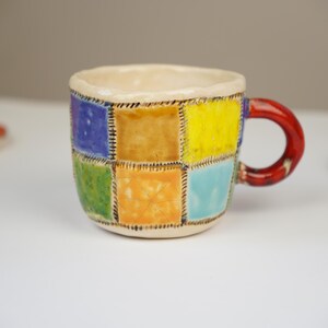 K&ouml;nnte beinhalten: Eine kleine Keramik-Kaffeetasse mit Patchwork-Design. Die Tasse ist mit Quadraten in verschiedenen Farben verziert, darunter Blau, Gr&uuml;n, Gelb und Orange. Der Henkel ist rot.