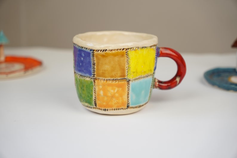 K&ouml;nnte beinhalten: Eine kleine Keramik-Kaffeetasse mit Patchwork-Design. Die Tasse ist mit Quadraten in verschiedenen Farben verziert, darunter Blau, Gr&uuml;n, Gelb und Orange. Der Henkel ist rot.