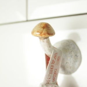 May include: A white ceramic hook shaped like a mushroom with a brown top. A white towel with a red label that says "100% cotton" is hanging on the hook.