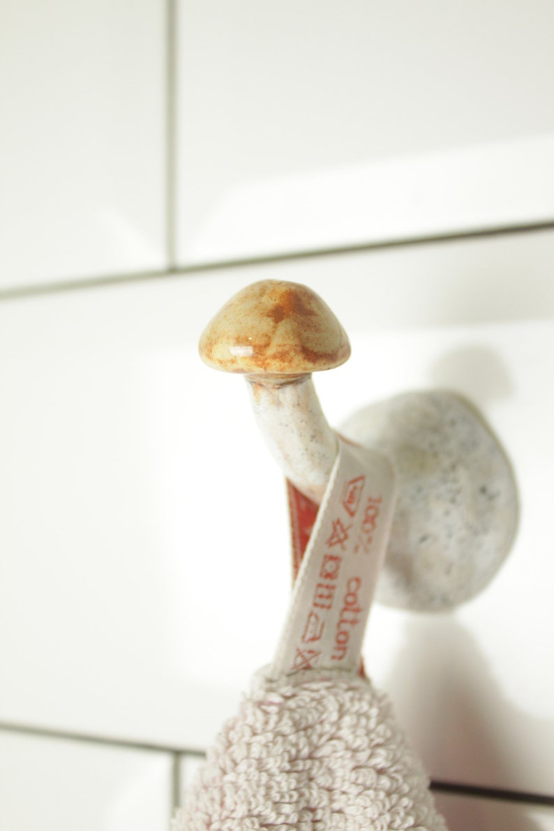May include: A white ceramic hook shaped like a mushroom with a brown top. A white towel with a red label that says "100% cotton" is hanging on the hook.