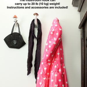 May include: Two white ceramic mushroom hooks mounted on a white wall. One hook has a black and white polka dot scarf hanging from it, and the other has a pink raincoat with white hearts hanging from it. A black face mask hangs from the left hook. The text on the wall reads: "Each hook has a secure, hidden metal bracket. The mount is suitable for any types of walls! (drywall, concrete, wood and other!) The mushroom hook can carry up to 20 lb (10 kg) weight! Instructions and accessories are included!"