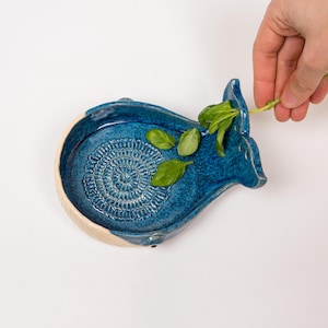 May include: A handcrafted, blue ceramic whale-shaped grater with a textured grating surface. The grater has a cream-colored rim and is decorated with fresh green basil leaves. A hand is placing more basil on the grater.