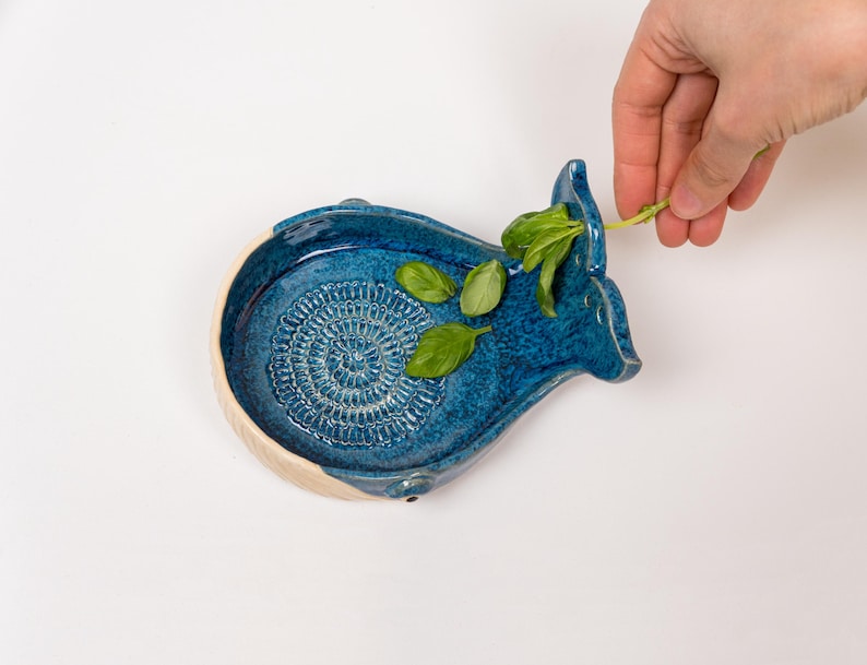 May include: A handcrafted, blue ceramic whale-shaped grater with a textured grating surface. The grater has a cream-colored rim and is decorated with fresh green basil leaves. A hand is placing more basil on the grater.
