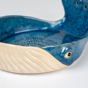 May include: A ceramic whale-shaped bowl with a blue interior and a cream-colored exterior. The bowl features a detailed eye and tail fin, and measures approximately 8 inches long.