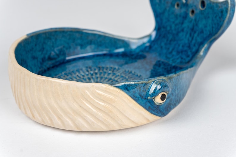 May include: A ceramic whale-shaped bowl with a blue interior and a cream-colored exterior. The bowl features a detailed eye and tail fin, and measures approximately 8 inches long.