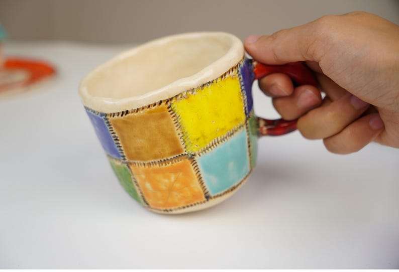 May include: A ceramic mug with a patchwork design. The mug is decorated with squares of yellow, orange, green, blue, and purple. The mug has a red handle.