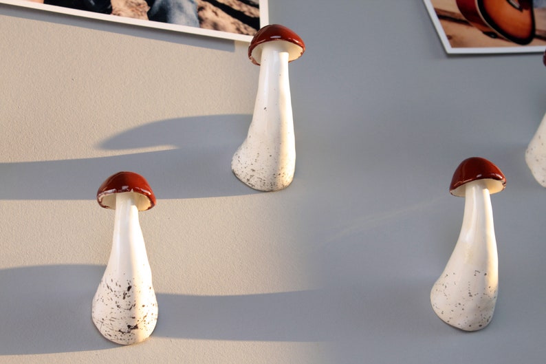 May include: Four ceramic mushroom-shaped hooks in white with brown tops. The hooks are mounted on a light gray wall.