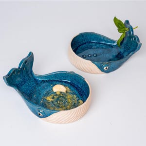 May include: Two ceramic whale-shaped dishes. The dishes are glazed in a deep blue with a textured interior and a cream-colored, ribbed base. One dish contains a small garnish. The whale's eye is a small, round detail.