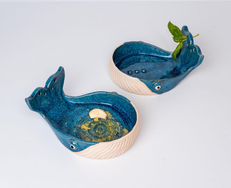 May include: Two ceramic whale-shaped dishes. The dishes are glazed in a deep blue with a textured interior and a cream-colored, ribbed base. One dish contains a small garnish. The whale's eye is a small, round detail.