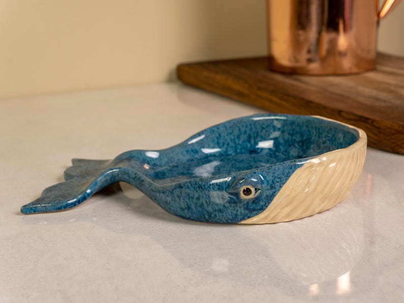 May include: A blue and white ceramic whale-shaped dish. The dish is glazed in a speckled blue and has a white belly with a sculpted whale eye.