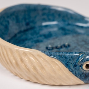May include: A ceramic bowl shaped like a whale with a blue and white glaze. The whale's tail is textured and the bowl has a smooth, rounded interior.