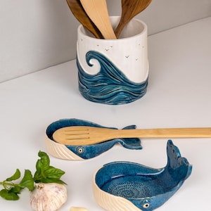 May include: Ceramic kitchenware set featuring a whale-shaped spoon rest and a utensil holder. The spoon rest is blue and white, with a whale tail design. The utensil holder has a wave design and holds wooden spoons.