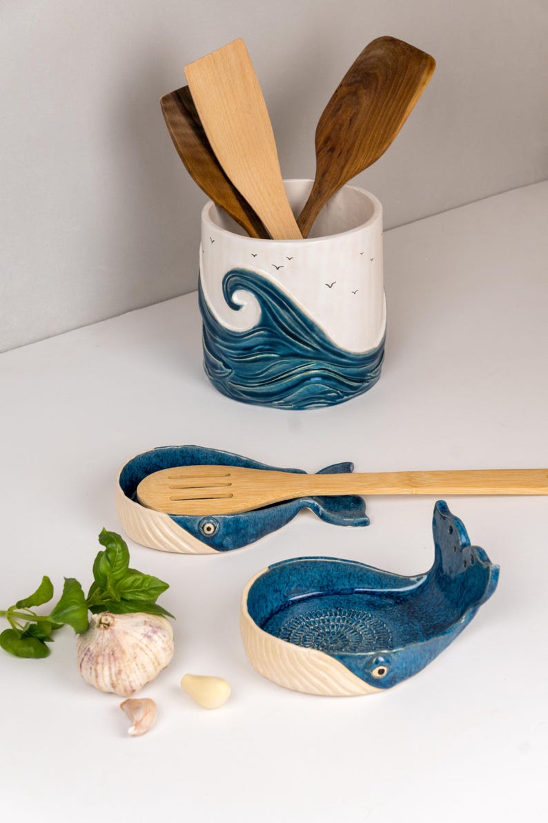 May include: Ceramic kitchenware set featuring a whale-shaped spoon rest and a utensil holder. The spoon rest is blue and white, with a whale tail design. The utensil holder has a wave design and holds wooden spoons.