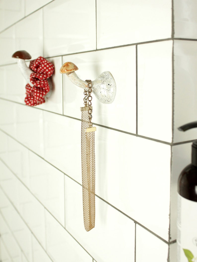 May include: Two ceramic mushroom hooks mounted on white subway tile. The hooks are white with brown tops and have a gold chain hanging from one.