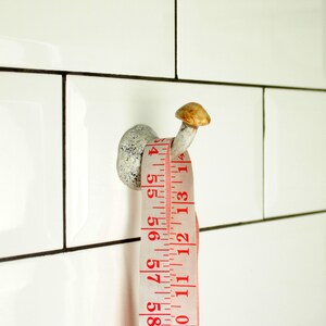 May include: A white ceramic hook with a mushroom-shaped top, with a white measuring tape wrapped around it. The tape has red numbers and markings, and the numbers visible are 6, 56, 57, 58, 59, 201, 5, 15, 4, 1314, 10, 11, 12, 13, 14.