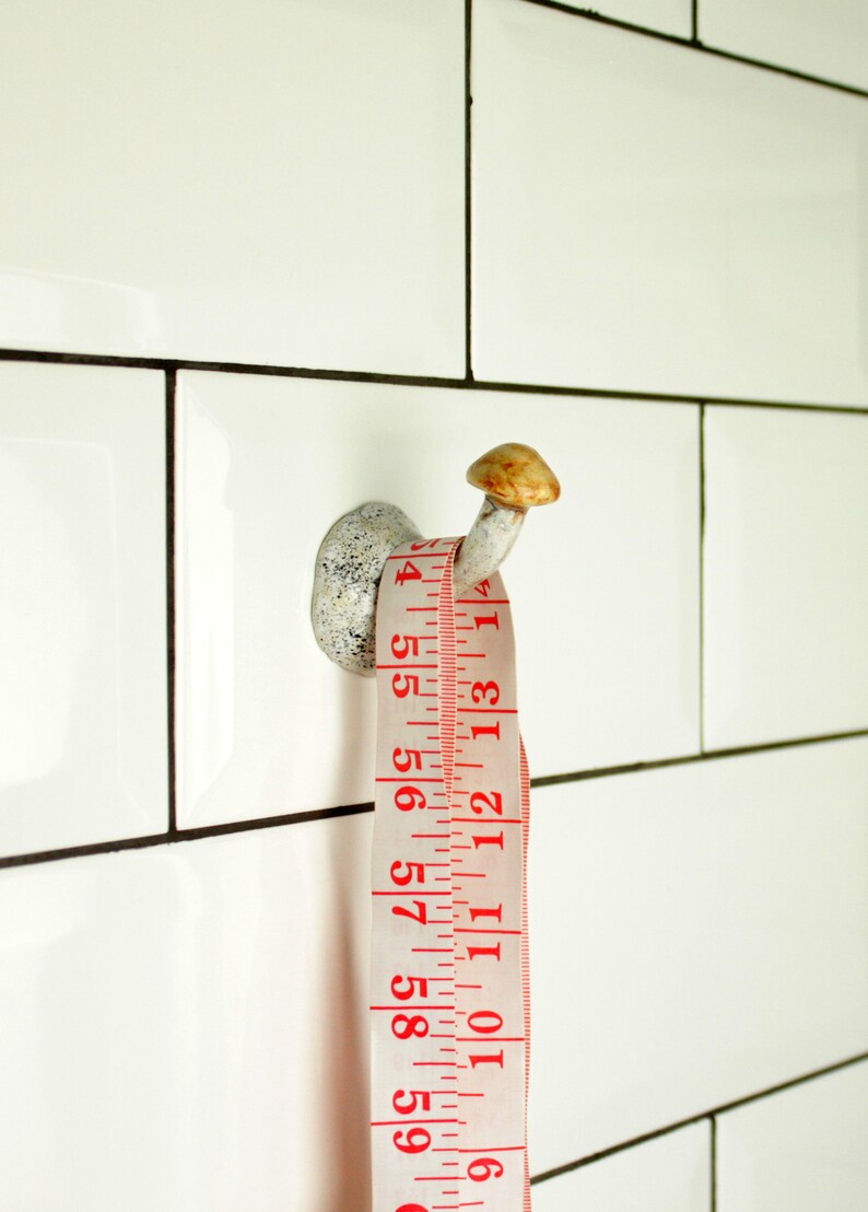 May include: A white ceramic hook with a mushroom-shaped top, with a white measuring tape wrapped around it. The tape has red numbers and markings, and the numbers visible are 6, 56, 57, 58, 59, 201, 5, 15, 4, 1314, 10, 11, 12, 13, 14.