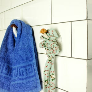 May include: A white tiled bathroom with a blue towel hanging on a mushroom-shaped hook and a green floral patterned scarf hanging on a mushroom-shaped hook. A bottle of hand soap labeled "BEAUTANICA" is on the counter.
