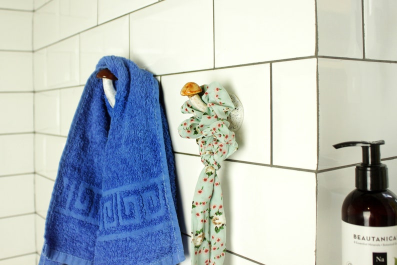 May include: A white tiled bathroom with a blue towel hanging on a mushroom-shaped hook and a green floral patterned scarf hanging on a mushroom-shaped hook. A bottle of hand soap labeled "BEAUTANICA" is on the counter.