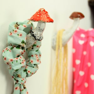 May include: Two ceramic mushroom hooks on a white wall. The hook on the left has a red and white mushroom top and a white stem. The hook on the right has a brown mushroom top and a white stem. A light blue scarf with a floral pattern is hanging on the left hook.