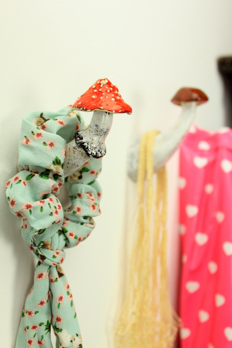 May include: Two ceramic mushroom hooks on a white wall. The hook on the left has a red and white mushroom top and a white stem. The hook on the right has a brown mushroom top and a white stem. A light blue scarf with a floral pattern is hanging on the left hook.