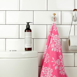 May include: A white bathroom with white subway tiles. A pink hand towel hangs on a mushroom-shaped hook. A roll of toilet paper hangs on a black cord from a mushroom-shaped hook. A brown bottle of liquid soap sits on a white toilet tank.